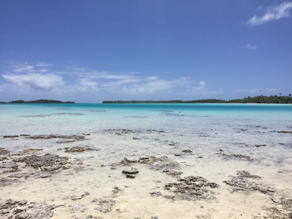 Vista della Laguna Blu di Rangiroa con acqua bassa e trasparente su sabbia bianca, rocce affioranti e mare dalle sfumature di azzurro, con il cielo che si fonde con l’orizzonte marino