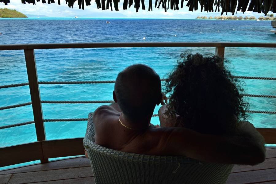 Coppia seduta e abbracciata su una poltrona del terrazzino del bungalow overwater, mentre guarda l'orizzonte sulla laguna di Bora Bora