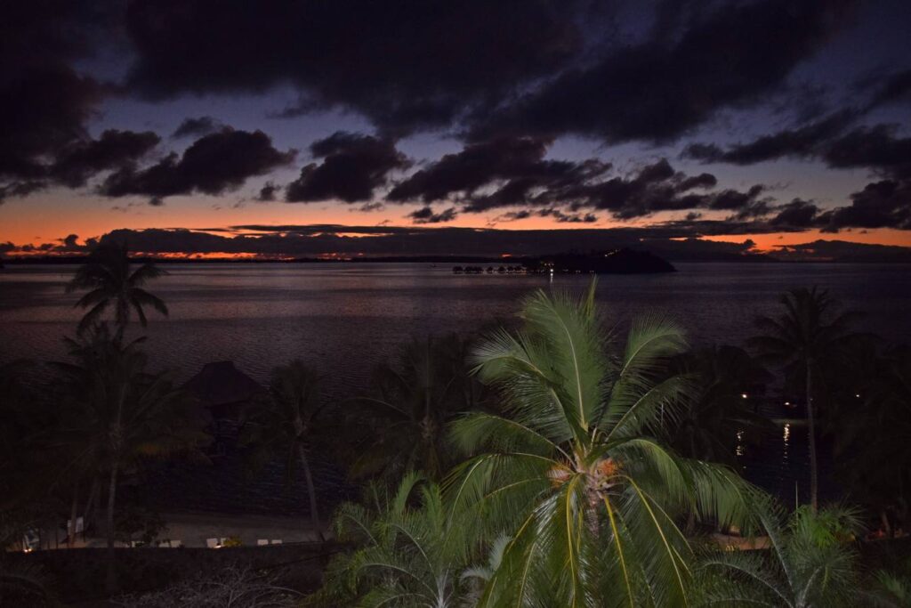 Vista dalla camera sulla laguna di Bora Bora tra il tramonto e la notte, con il mare, le palme e i bungalow overwater illuminati dalle ultime luci del cielo