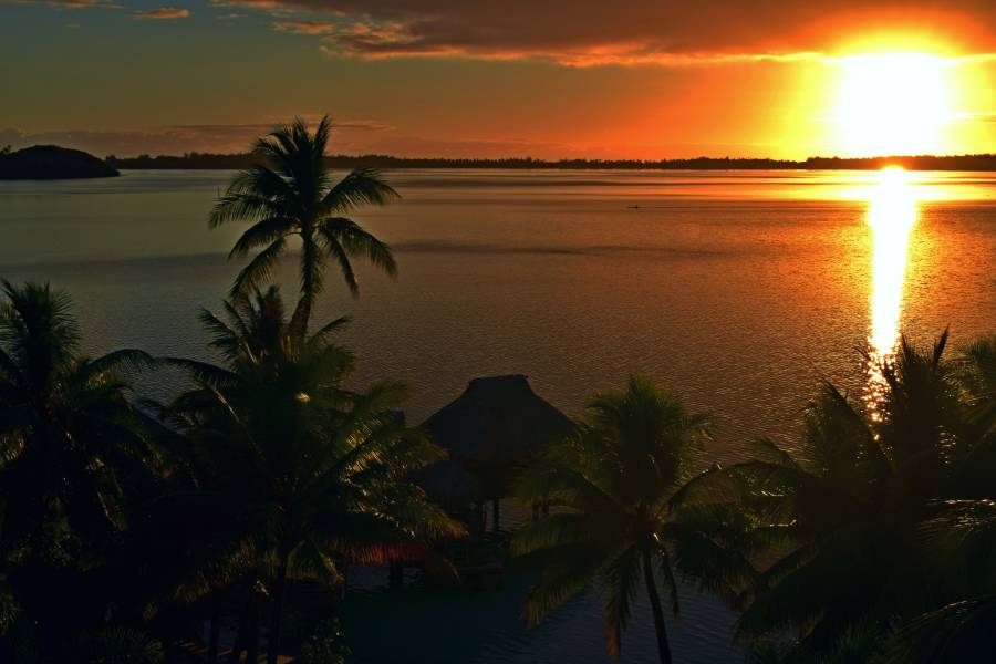Vista dalla camera sulla laguna di Bora Bora al tramonto, con il mare in primo piano, le palme e i bungalow overwater immersi nelle sfumature calde del sole